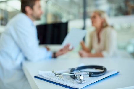 a stethoscope and a clipboard on a desk with a doctor and patient in the background